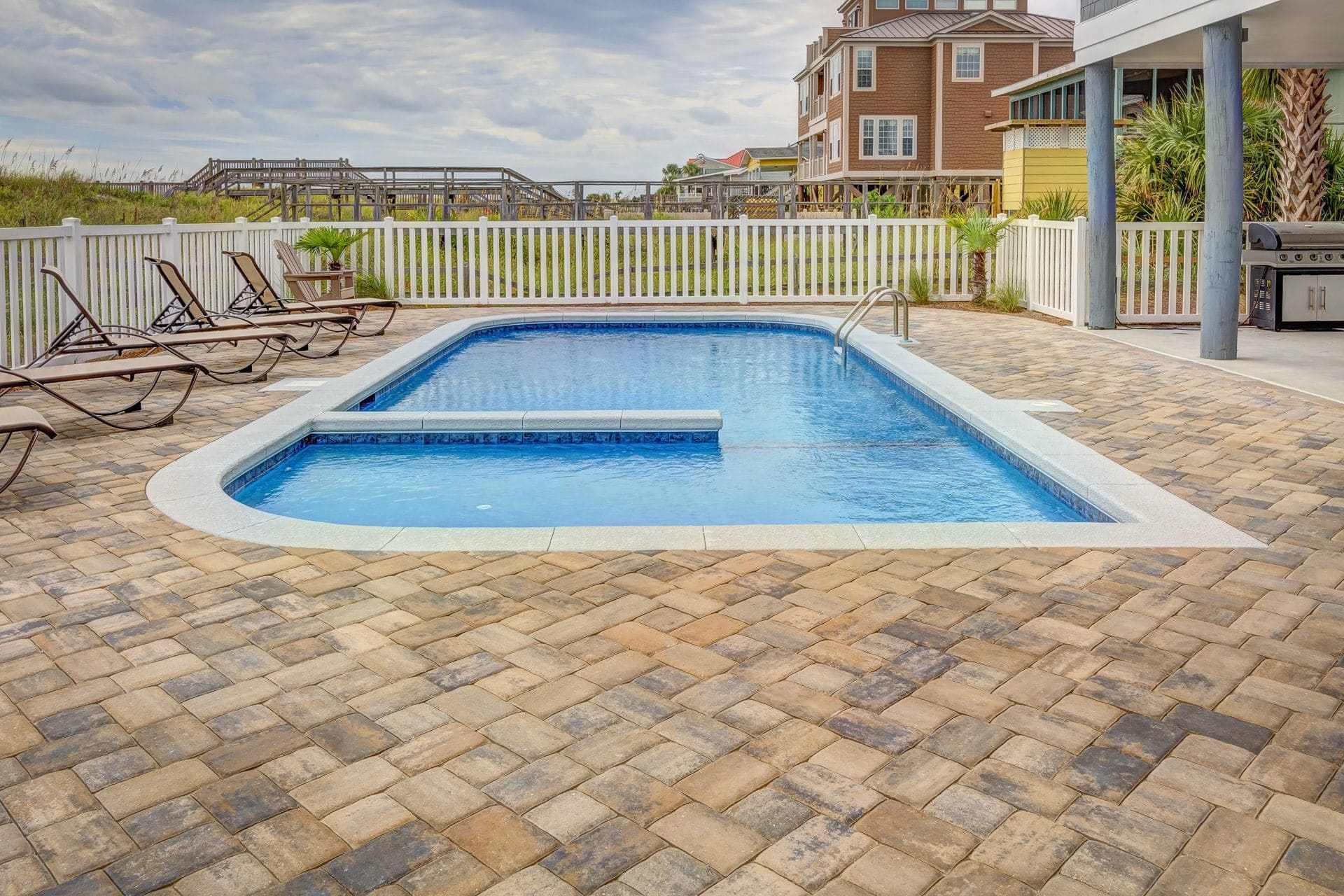 Inground backyard pool with a paved patio, lounge chairs and a white fence